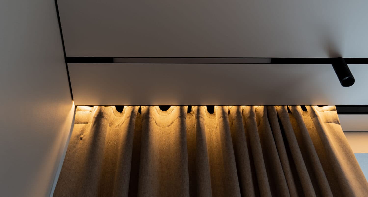 Detaliu cu nișă de draperie iluminată cald în tavan extensibil, cu șină magnetică neagră și spot cilindric
