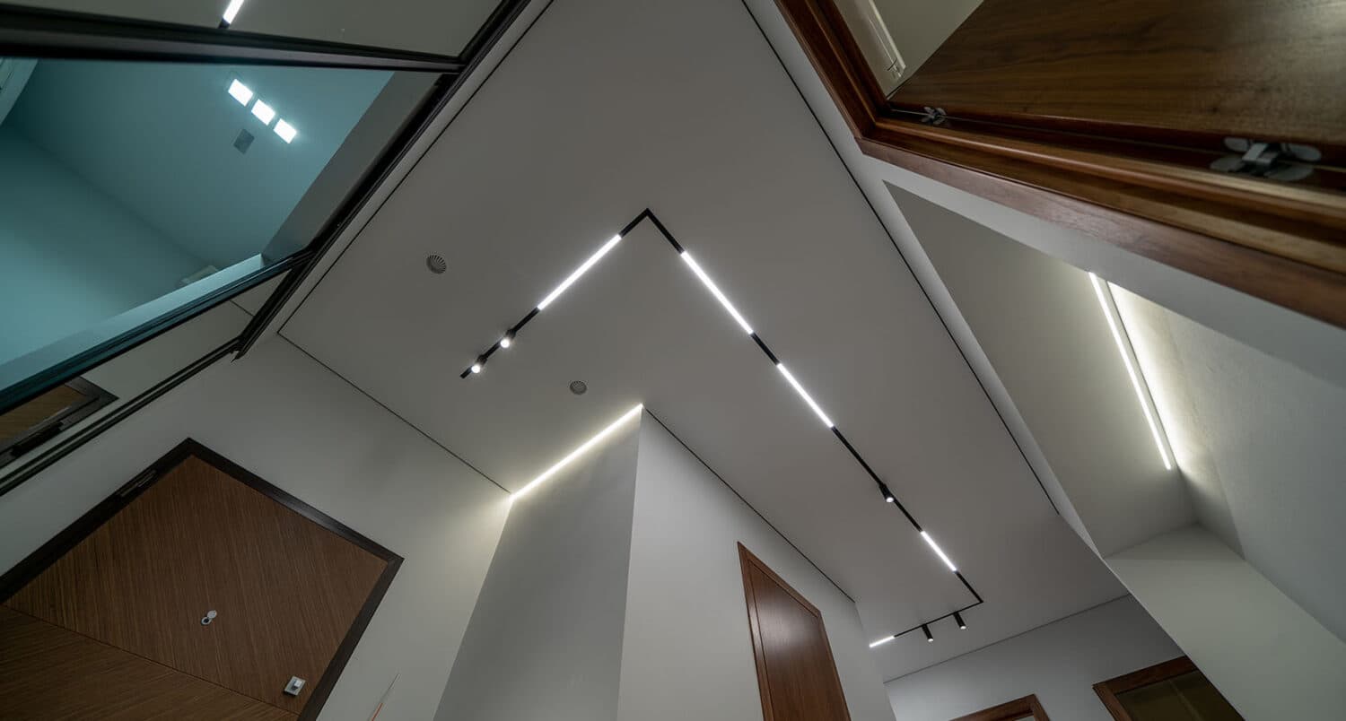 Trasee de șină magnetică cu segmente LED albe pe tavanul extensibil al unui hol cu uși și mobilier din nuc