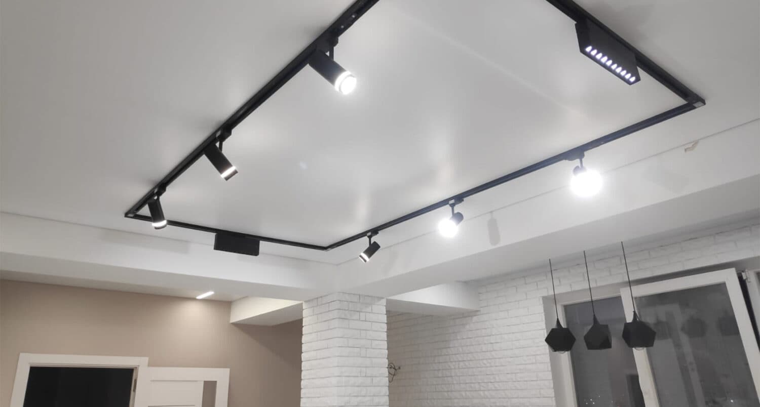 Cadru de șină magnetică neagră cu spoturi cilindrice pe tavan extensibil alb mat, într-un living cu pereți din cărămidă albă